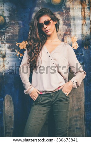 trendy fashion girl in silky beige shirt, green pants and sunglasses stand in front old rusty grunge metal door, outdoors