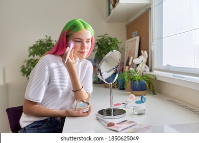 Trendy, fashionable teenager girl with dyed colored hair doing makeup looking in mirror with brush of decorative cosmetics sitting at table at home