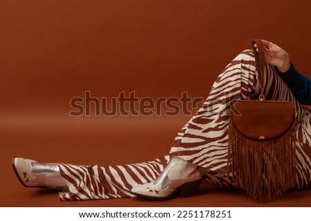 Trendy fashionable woman's outfit details: brown suede fringed bag, flared trousers with zebra print, white cowboy boots,. Copy, empty space for text