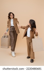 Trendy Girl With Shopping Bags Holding Hands With Cheerful And Stylish Mom On Beige Background