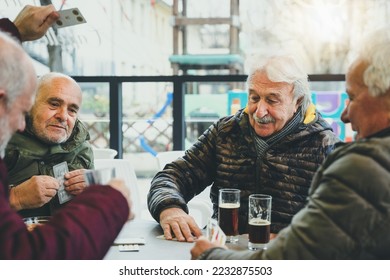 Trendy group of senior having fun at bar playing card at table with drinks-Cheerful old man drinking and enjoying free time together at patio restaurant-Focus on central men's-Friendship Games Concept - Powered by Shutterstock