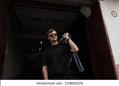 Trendy Handsome Young Man Hipster Posing In A Dark Warehouse. Modern Cool Guy In Sunglasses In Fashionable Clothes With A Stylish Hairstyle With A Brutal Beard In A Dark Room. Youth Menswear.