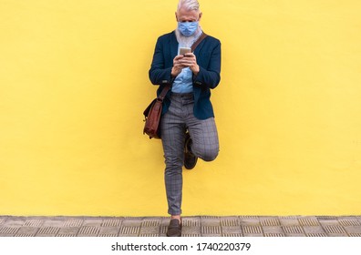 Trendy hipster man using mobile phone outdoor while wearing fashion silk safety mask with yellow wall background - Outfit, technology and lifestyle during coronavirus time - Focus on hands - Powered by Shutterstock