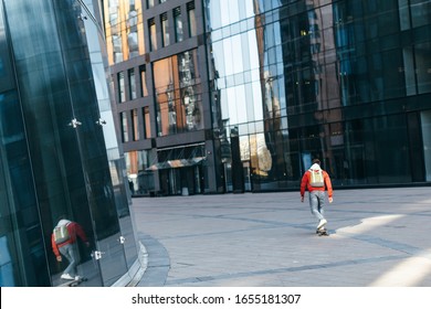 Trendy hipster in red jacket with green backpack riding on longboard among the modern houses. Urban and street photo. Healthy lifestyle, fashion look. Concept of leisure activity and youth.