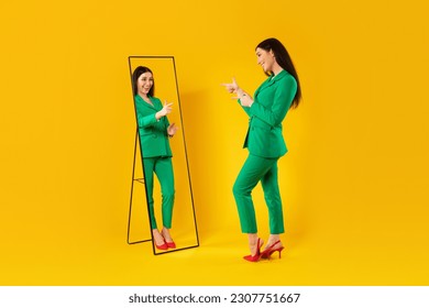 Trendy lady pointing at her reflection in mirror posing after successful shopping, wearing green jacket trouser suit, standing on yellow studio background. I like this outfit concept