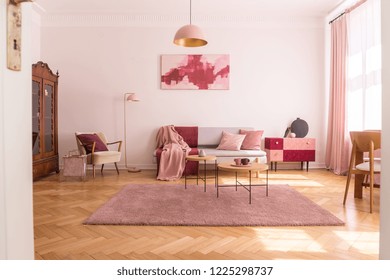 Trendy living room interior with grey couch with pastel pink pillows and blanket, stylish beige armchair with burgundy pillow and retro cabinet in the corner