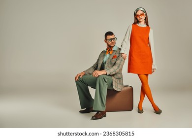 trendy man sitting on vintage suitcase near woman in orange dress on grey, retro-inspired couple - Powered by Shutterstock