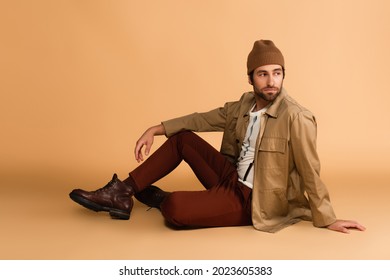 trendy man in stylish autumn outfit looking away while sitting on beige - Powered by Shutterstock