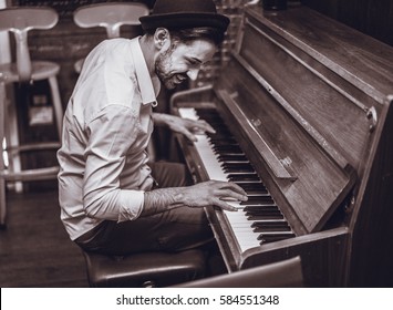 Trendy Man With Stylish Hat And Beard Trying Playing Vintage Old Wooden Piano - Young Artist Performing In Cocktail Bar - Black And White Editing - Focus On Ear Eye - Warm Contrast Filter