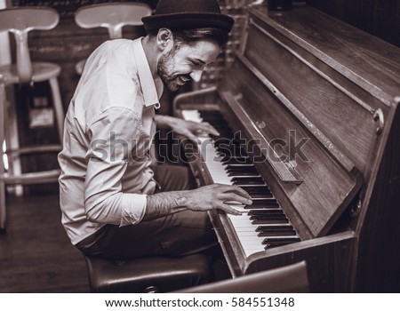 Trendy man with stylish hat and beard trying playing vintage old wooden piano - Young artist performing in cocktail bar - Black and white editing - Focus on ear eye - Warm contrast filter