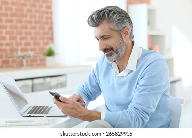 Trendy Mature Man Using Smartphone In Front Of Laptop