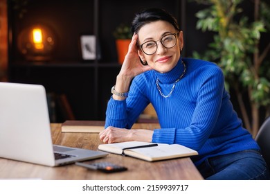 Trendy middle aged woman in blue turtleneck and glasses smiling and  looking at camera  while sitting at table and resting during break in remote work in home office