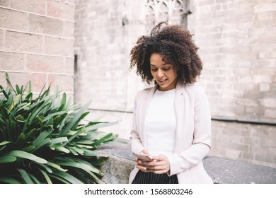 Trendy Millennial African American Female In Formal Clothes Focusing On Screen And Using Smartphone While Standing On Street Against Shabby Stone Building In Gothic Style In Barcelona