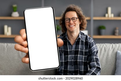 Trendy Mobile Phone. Happy Curly Male In Glasses Holding Smartphone With White Empty Screen In Hand, Showing Device Close To Camera, Sitting On Couch At Home. Gadget With Blank Space, Mock Up