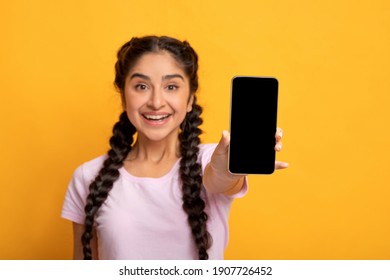 Trendy Mobile Phone. Portrait of cheerful young indian woman holding cellphone with black blank screen in hand, showing device. Gadget with empty free space for mock up, selective focus, blur