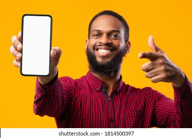 Trendy Mobile Phone. Smiling bearded black man holding latest slim smartphone with white blank screen in hand and pointing at it, indicating empty space for mock up, blurred background
