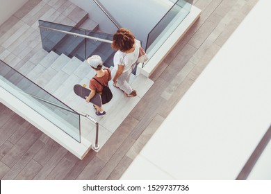 Trendy mother and son walking on stairs in a modern city context. Young boy with skateboard goes down the ladder with mum Top view of people walks in a new mall. Family scene at the shopping center - Powered by Shutterstock