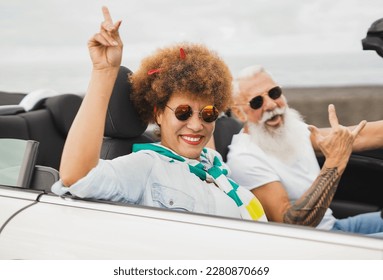 Trendy mulitracial senior couple having fun dancing inside convertible car - Mature people on a road trip in summer vacation with cabriolet car - Powered by Shutterstock