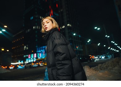 Trendy Night Portrait Of Stylish Woman In Black Winter Jacket Posing At Camera With Serious Face On Cityscape Background.