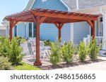 Trendy outdoor patio pergola shade structure made of dark wood, awning and patio roof, white dining table, white chairs, metal grill surrounded by landscaping in a luxury American neighborhood