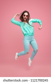 Trendy Rebel Woman In Blue Sportive Outfit And Sunglasses Jumping Up And Screaming On Pink Background 