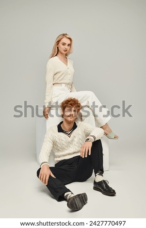 trendy redhead man looking at camera near blonde woman sitting on white cube on grey, classic style