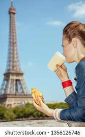 Trendy Solo Traveller Woman In Blue Jeans Overall Not Far From Eiffel Tower In Paris, France Having Coffee Cup And Croissant.