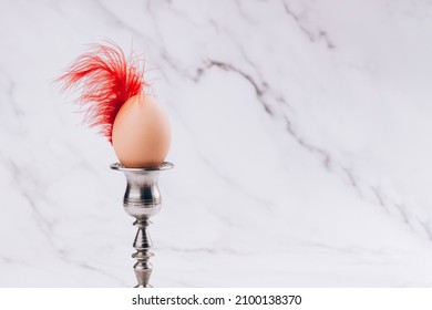 Trendy still life vertical Easter composition with hen egg in vintage candlestick with bright red feather against light marble background. Easter egg as a symbol of rebirth and circle of life