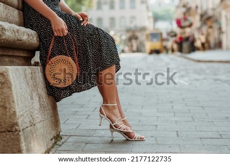 Trendy summer rotan wicker bag, white strap sandals in stylish female outfit. Woman posing in street of European city. Fashion details. Copy, empty space for text