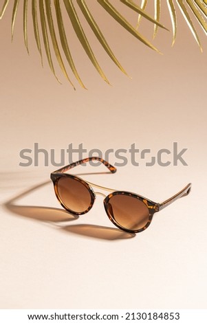 Trendy sunglasses still life in minimal stile. Womens sunglasses on a beige background with golden palm leaf - summer fashion concept. Fashionable accessories. Optic store discount, sale. Vertical