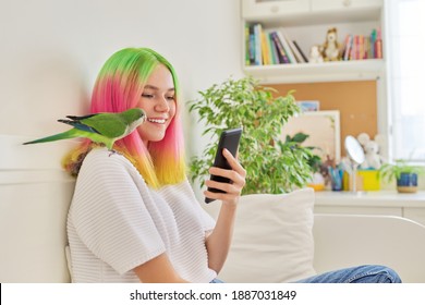 Trendy Teen Girl With Smartphone And Young Green Quaker Parrot On Her Shoulder, At Home On Bed. Pet Owner Talking On Video Communication, Human Bird Friendship, Lifestyle, Communication