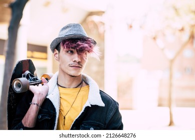 Trendy Teenager Boy Portrait Holding Skate Board And Wearing Hat. Diversity Teen People With Coloured Violet Hair And Moustaches Looking On His Side And Walking In The City