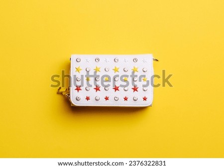 Trendy white leather women's purse covered with red, yellow, and white rivets-stars and rhinestones on a vibrant yellow background. Creative mockup for advertising a handbag store. Fashion trend.