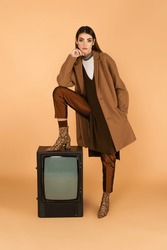 Trendy Woman With Hand In Pocket Of Trendy Coat Stepping On Vintage Tv On Beige