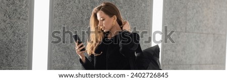Trendy woman in jacket with backpack using cellphone while standing near grey concrete wall, banner