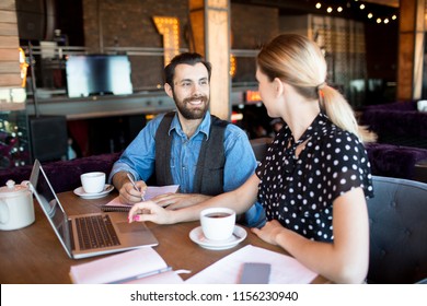 Trendy woman and man sitting with laptop and teacups at table chatting and working in relaxation 
