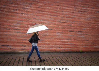 Trendy Woman With Umbrella Is Walking Next To Red Brick Wall In Rain. Side View Of Woman Wearing Casual Clothing And Face Mask Walk At Pedestrian Sidewalk. Rainy Season In City