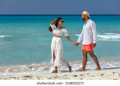 Trendy young couple in love on a tropical beach walking along the edge of the ocean hand in hand smiling into each others eyes on a hot sunny day.