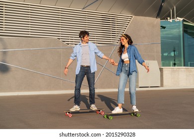 Trendy young couple riding longboards together holding hands. Stylish caucasian man and woman wearing street fashion clothes longboarding in summer city urban space. Happy casual friends on skateboard