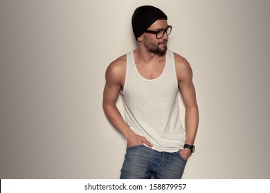 Trendy young man wearing blue jeans, a tank top and a beanie hat leaning against a wall
