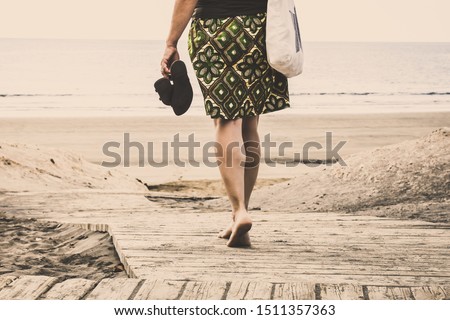 Trendy young woman, walking barefoot on a rustic wooden walk towards the beach and the sea. Female legs back view of a girl walking with leather sandals in hand. Summer vacation and holiday concept.
