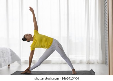 Triangle pose (yoga posture, Trikonasana) Middle-aged Asian woman doing yoga on one hand touching the calf the other hand holds up the ear. Bedroom background. - Powered by Shutterstock