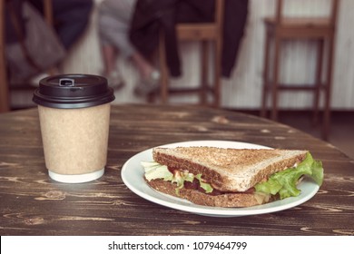 Triangle Sandwich And Takeaway Coffee Cup Of Craft Paper On Wooden Table, With Sitting People On Background