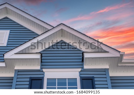 Triangle shape decorative gable with colonial white soffit and fascia on a blue horizontal vinyl siding modern American estate home with colorful dramatic orange sunset sky