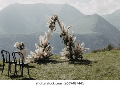A triangular boho wedding arch with pampas grass and pastel flowers on a grassy hill in Gudauri, Georgia, framed by Caucasus mountains under a clear sky.