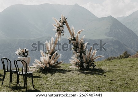 A triangular boho wedding arch with pampas grass and pastel flowers on a grassy hill in Gudauri, Georgia, framed by Caucasus mountains under a clear sky.
