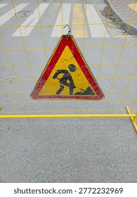 Triangular construction warning sign hanging on a yellow barrier fence near a pedestrian crossing on an urban street, signaling road repairs and safety precautions.
