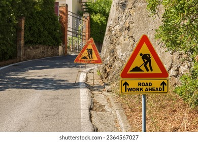 Triangular orange, red and black road work ahead sign or work in progress sign, by the side of a road under maintenance (bad condition).