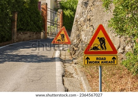 Triangular orange, red and black road work ahead sign or work in progress sign, by the side of a road under maintenance (bad condition).