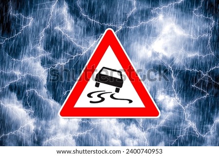 A triangular pictogram in the shape of a warning triangle with a danger warning of slippery roads and extreme weather in front of a dramatic sky with flashing lightning and heavy rain